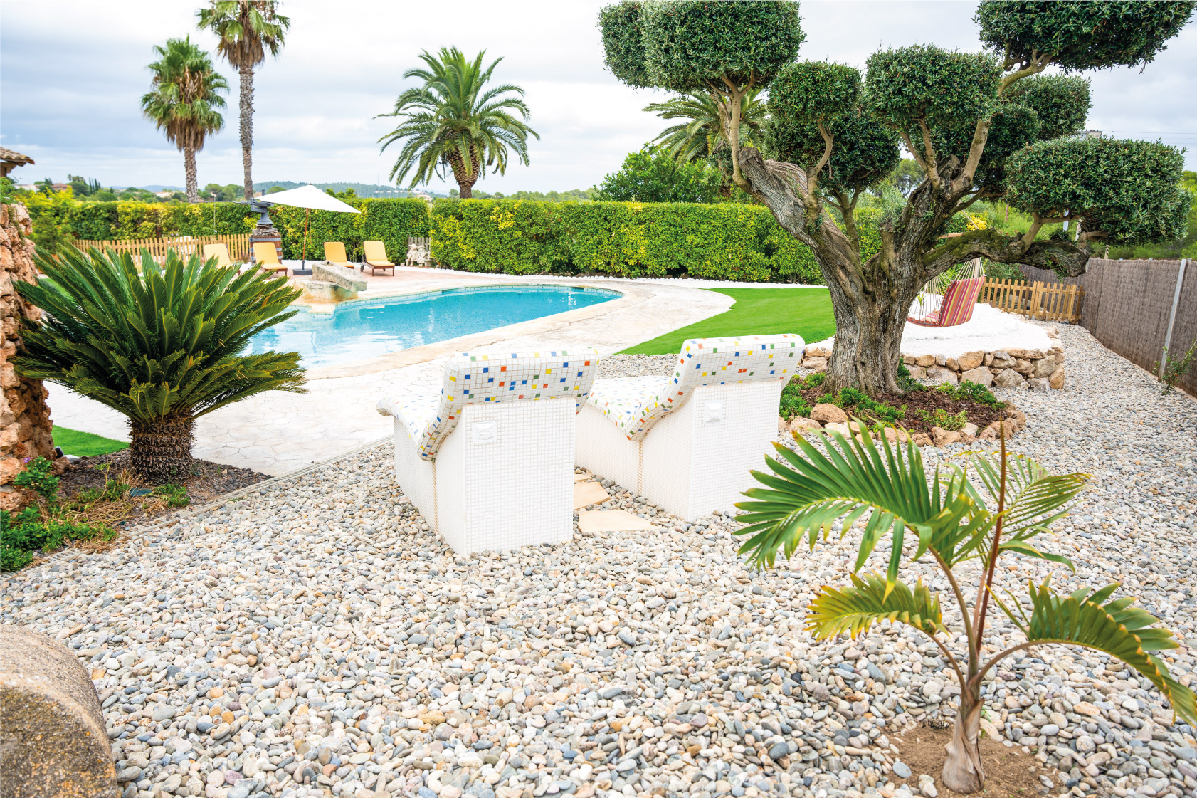 Butaques de mosaic davant la piscina de la Masia L’Encís, envoltades de jardí i vegetació mediterrània