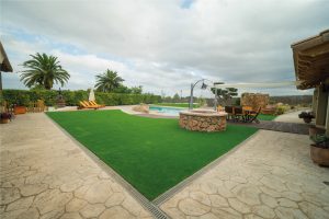 Zona enjardinada amb gespa i piscina a la Masia L’Encís, amb taula exterior i sínia decorativa de pedra