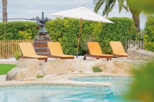 Zona de relax de la Masia L’Encís amb tumbones, para-sol i font antiga al costat de la piscina