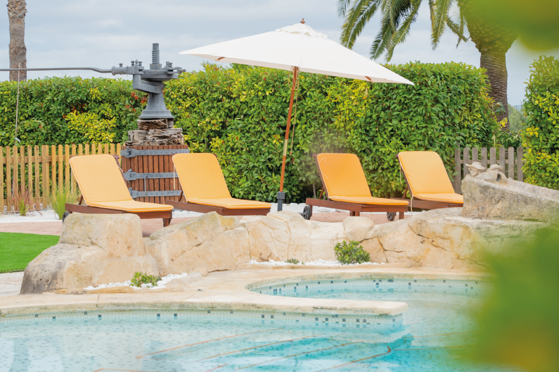 Zona de relax de la Masia L’Encís amb tumbones, para-sol i font antiga al costat de la piscina