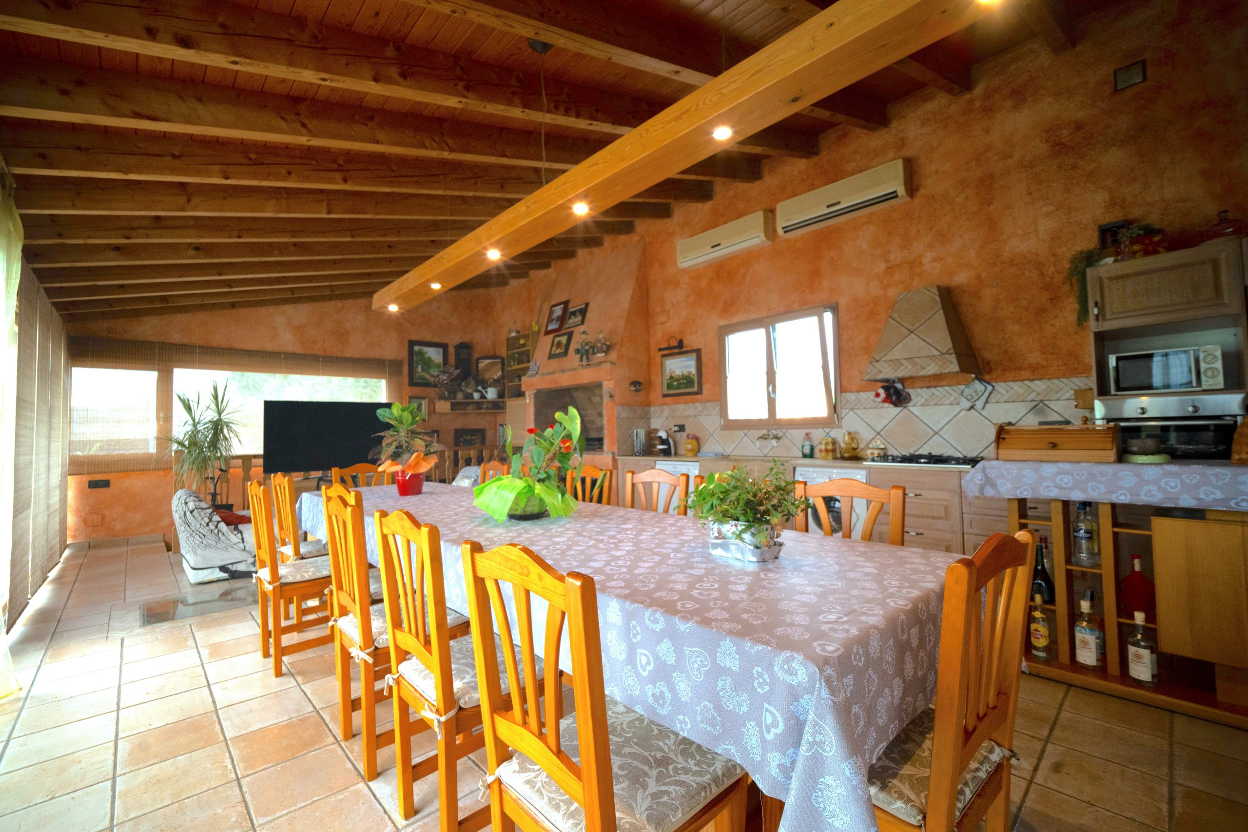 Menjador rústic interior amb cuina, xemeneia i taula llarga a la Masia L’Encís