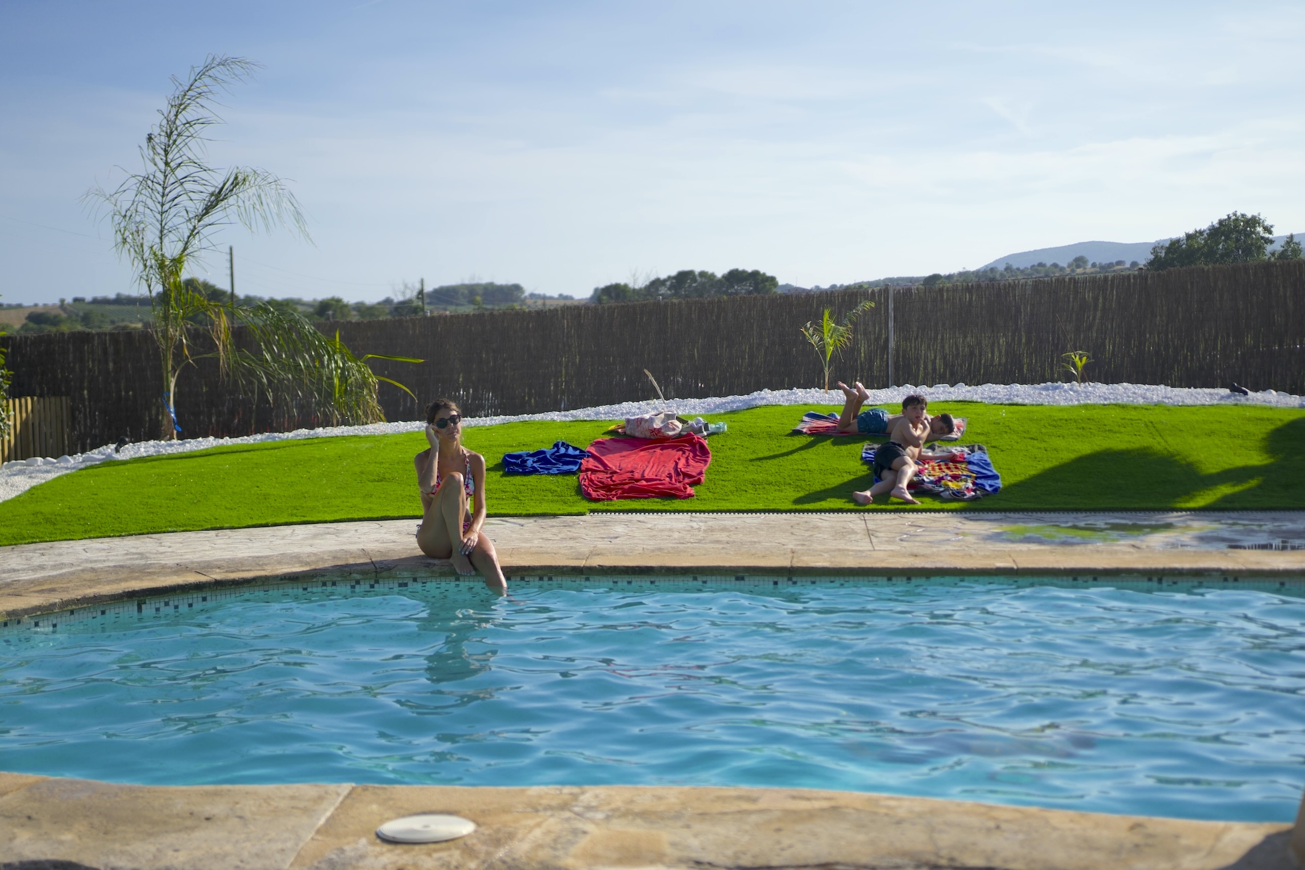 Una dona relaxant-se a la vora de la piscina mentre una família descansa al jardí de la Masia L’Encís
